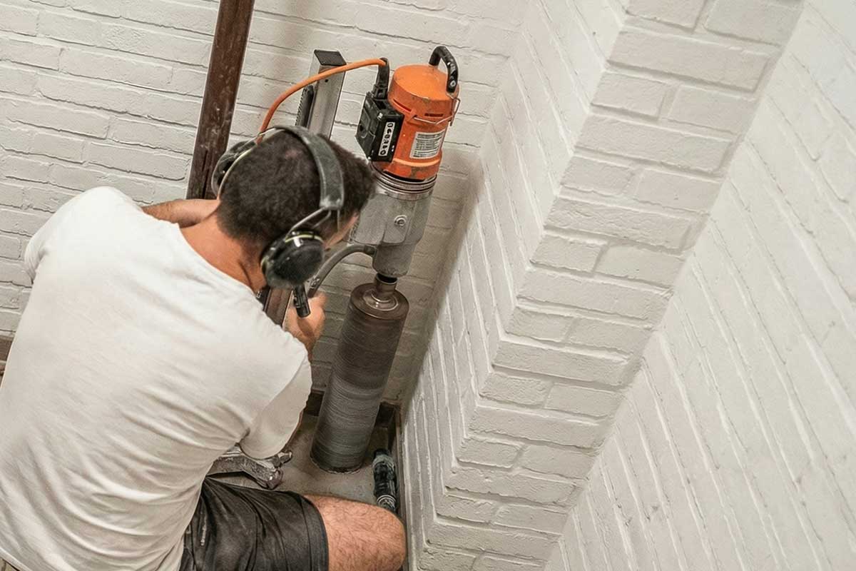 Arbeiter mit Gehörschutz beim Kernbohren durch weiß gestrichenes Ziegelmauerwerk